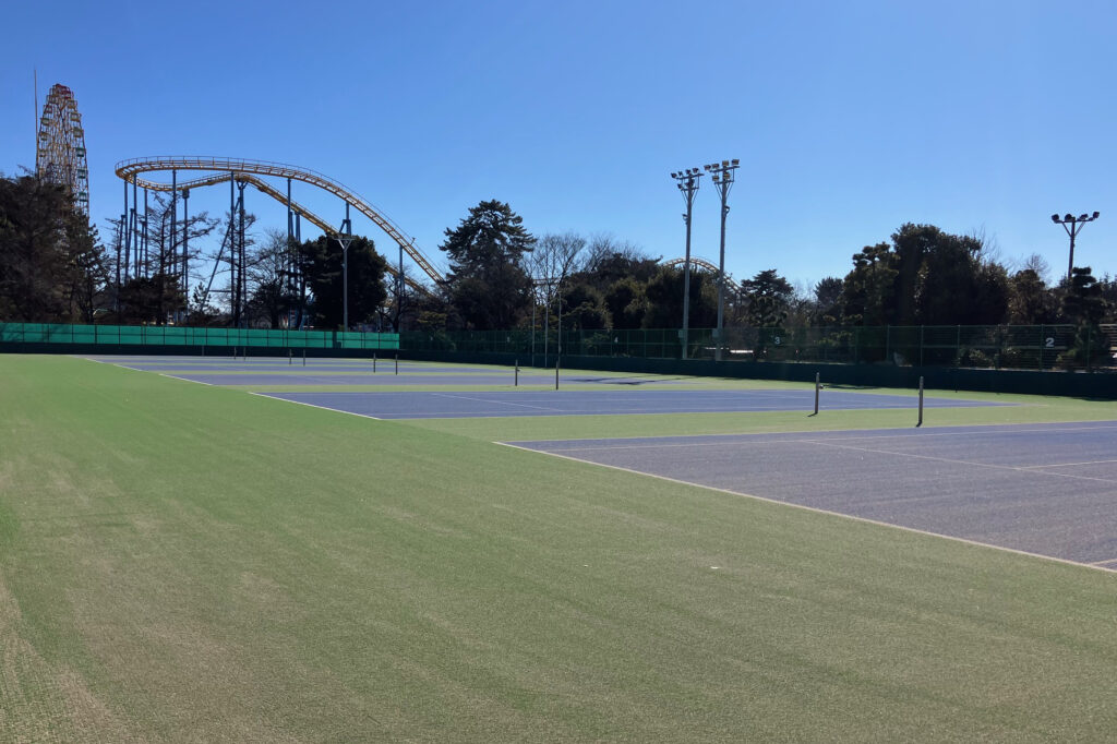グリーンとブルーのよるカラーコーディネーションのテニスコート、奥に華蔵寺遊園地のジェットコースターが見える